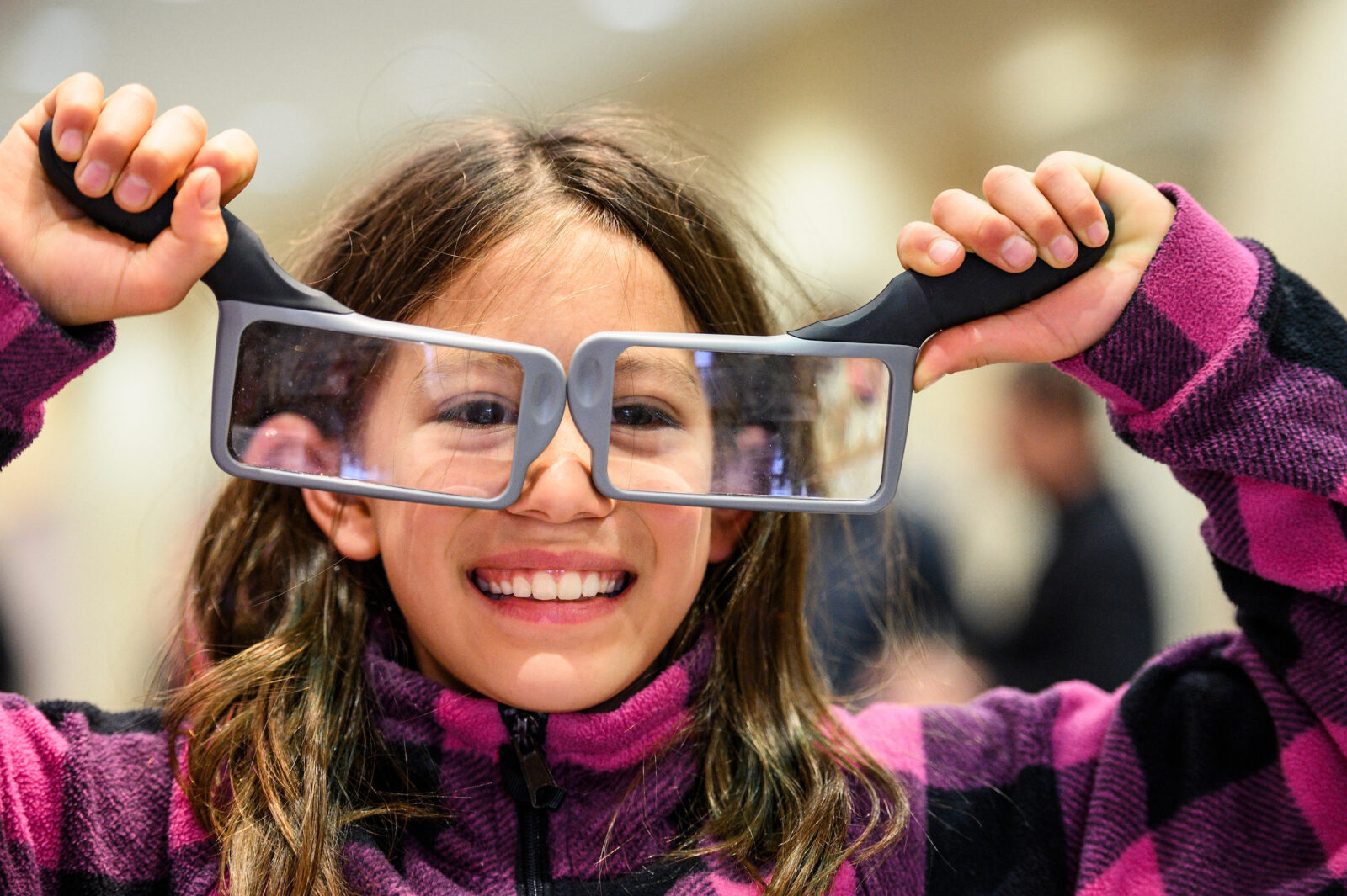 A young girl with long brown hair is smiling as she holds up an optical device that looks like large glasses to her face