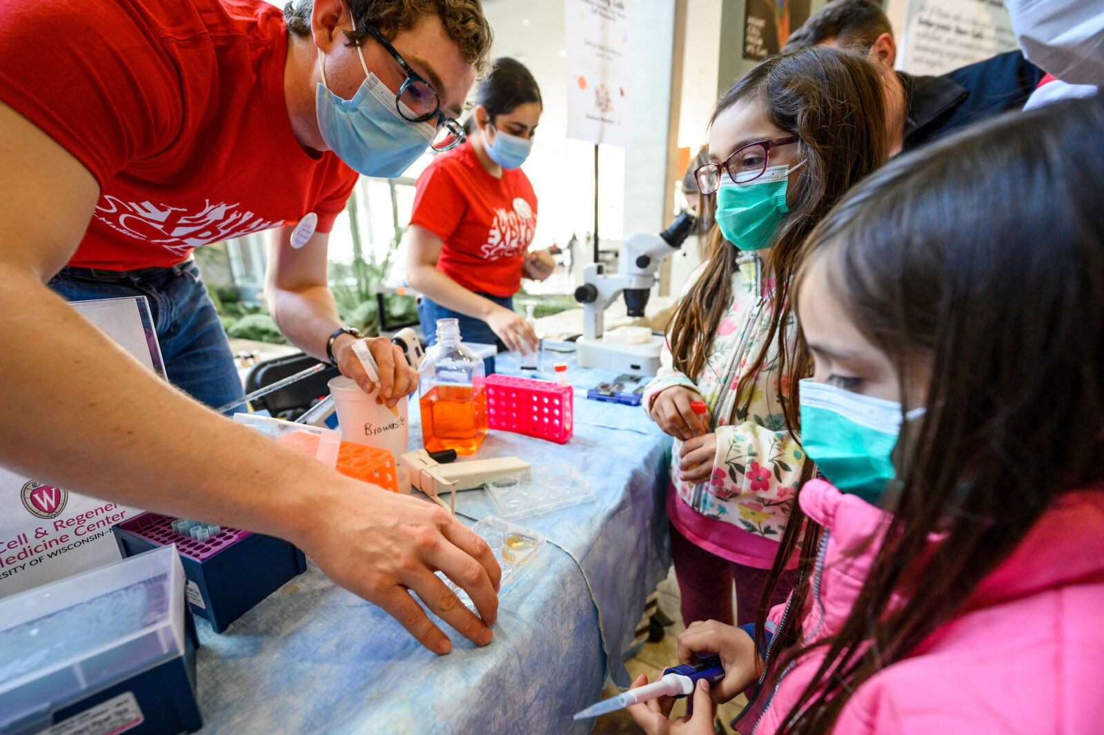 At left, undergraduate student and Goldwater Scholar Samuel Neumann helps 8-year-old Maria Escalona (center) and 6-year-old Ignacio Escalona (right) feed and pipet mock cell samples